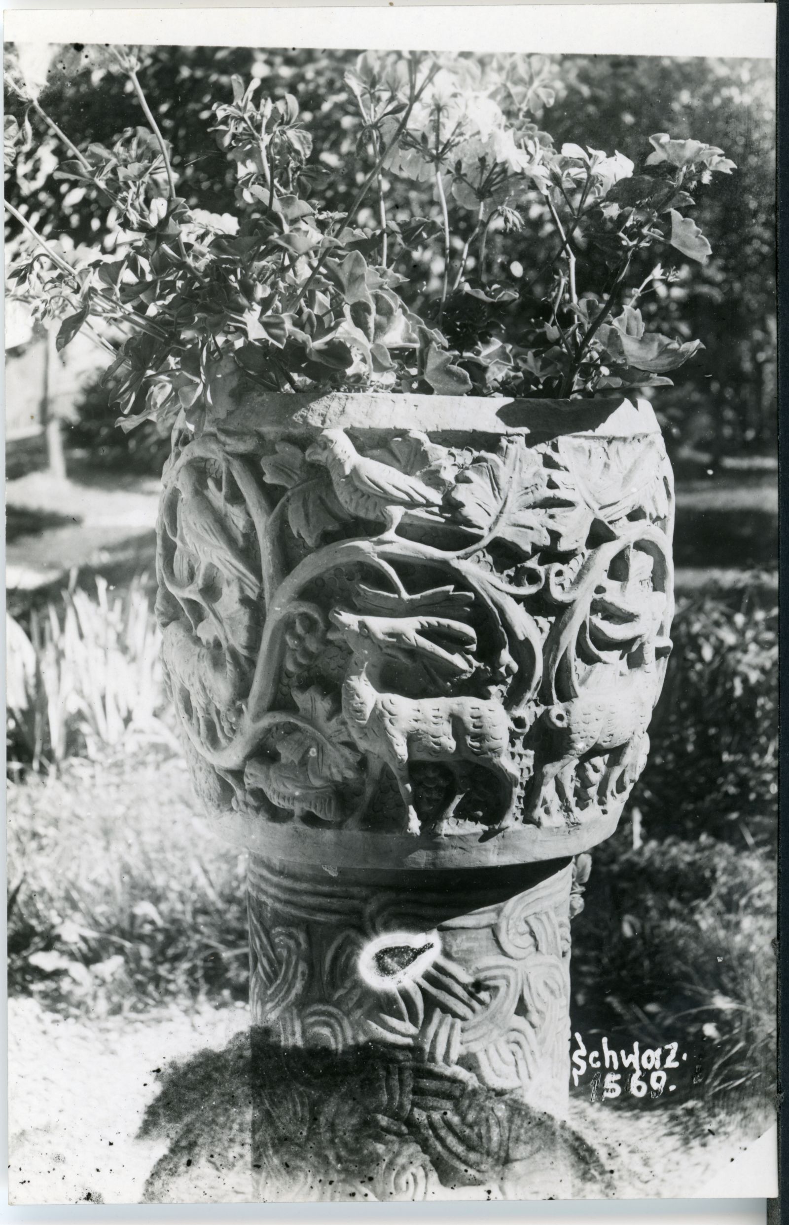 Steinvase im Garten mit Blumen bepflanzt. Flechtwerk in romanischem Stil am Sockel und gotisches Rankenwerk mit Tieren am Gefäß. Historismus.